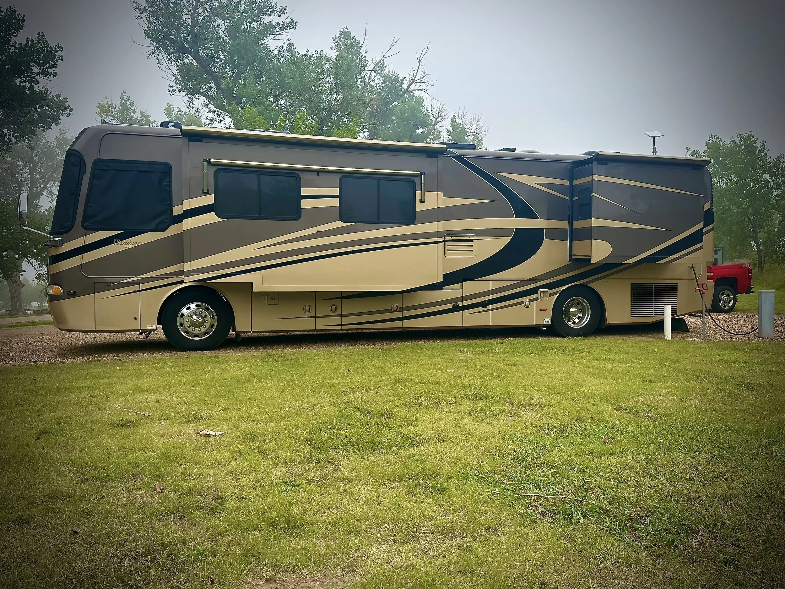 A beige and black RV parked on a grassy area with trees in the background, cloudy sky overhead.
