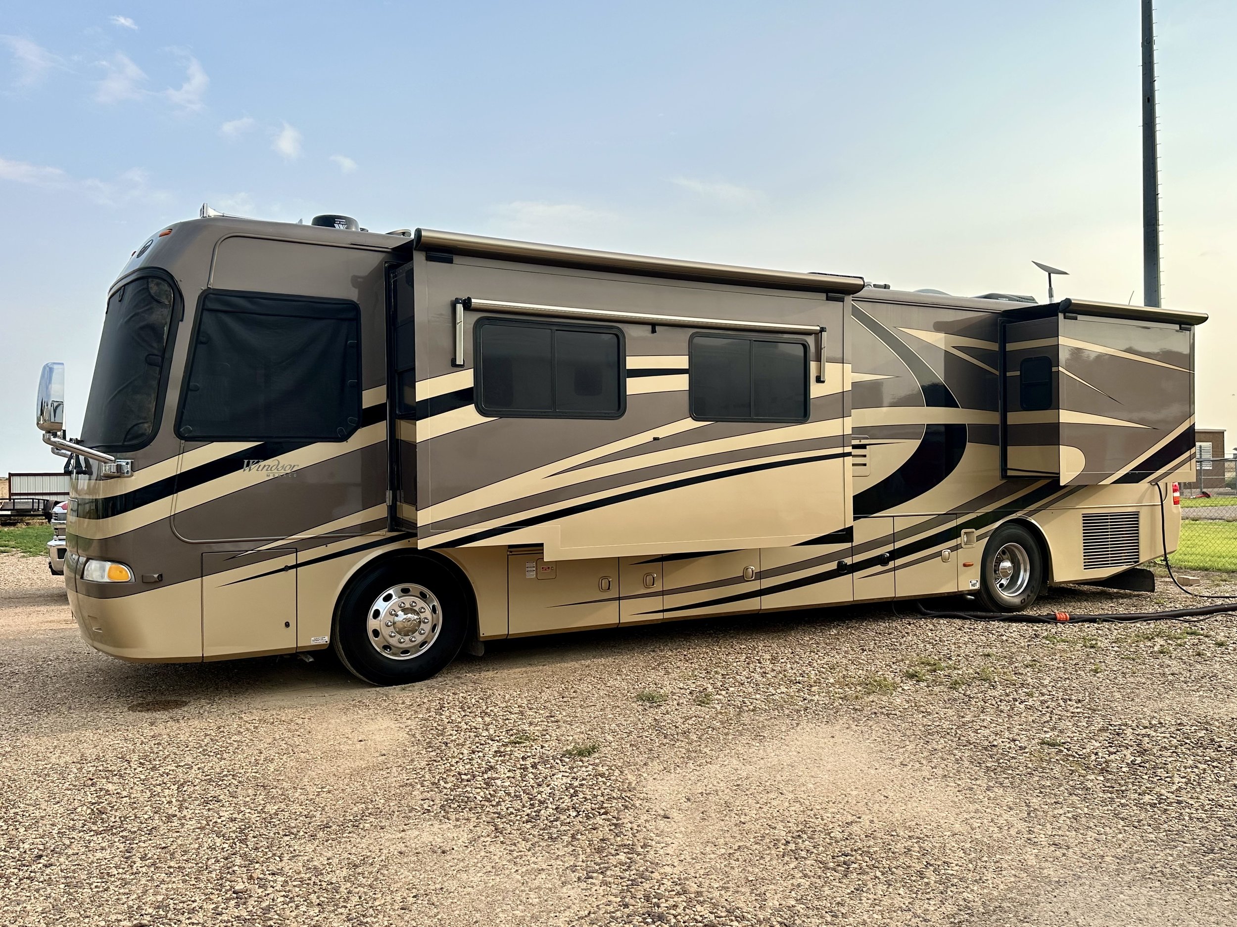 A beige and black travel trailer with multiple windows and a slide-out section, parked on a gravel lot, under a partly cloudy sky.