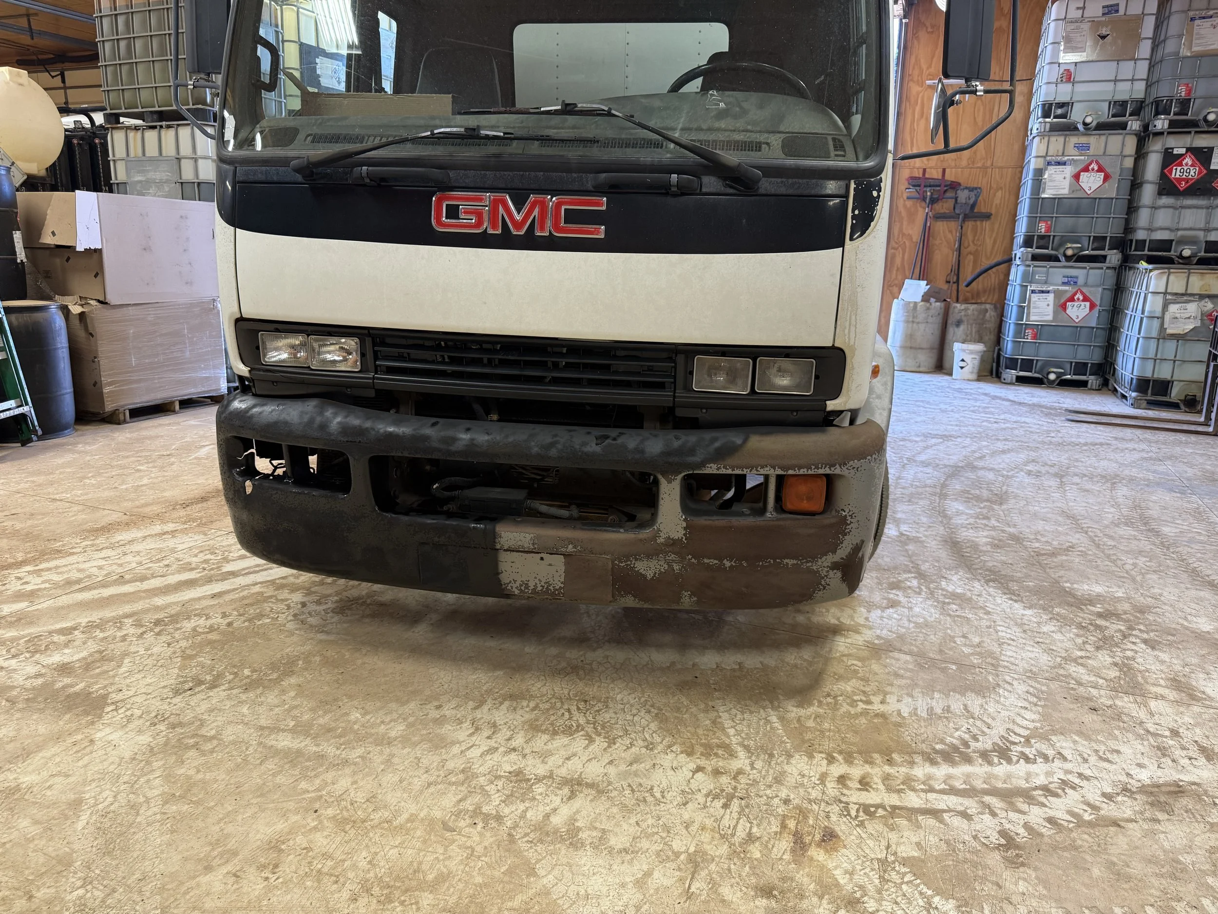 Front view of a white GMC truck parked inside a storage or warehouse area with various containers and equipment around.