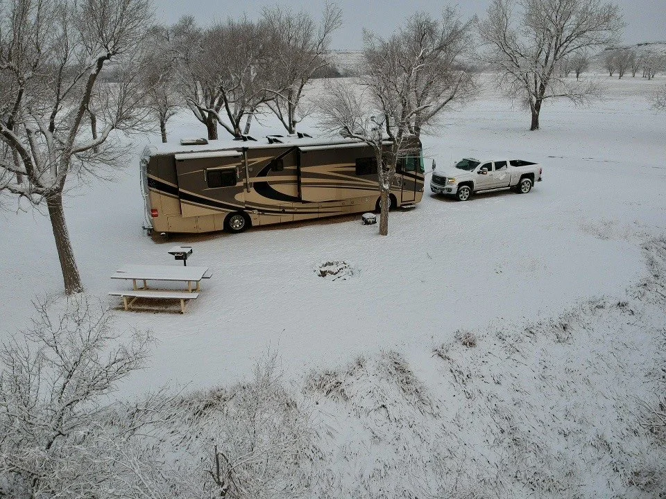 A motorhome and pickup truck parked in a snowy field with leafless trees, a picnic table, and a fire pit. Cold Winter