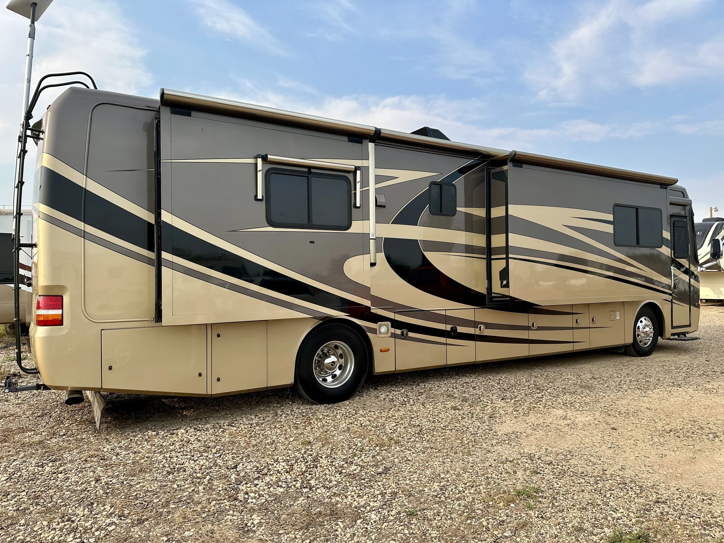 A large modern recreational vehicle (RV) parked on gravel with a blue sky and some clouds above.