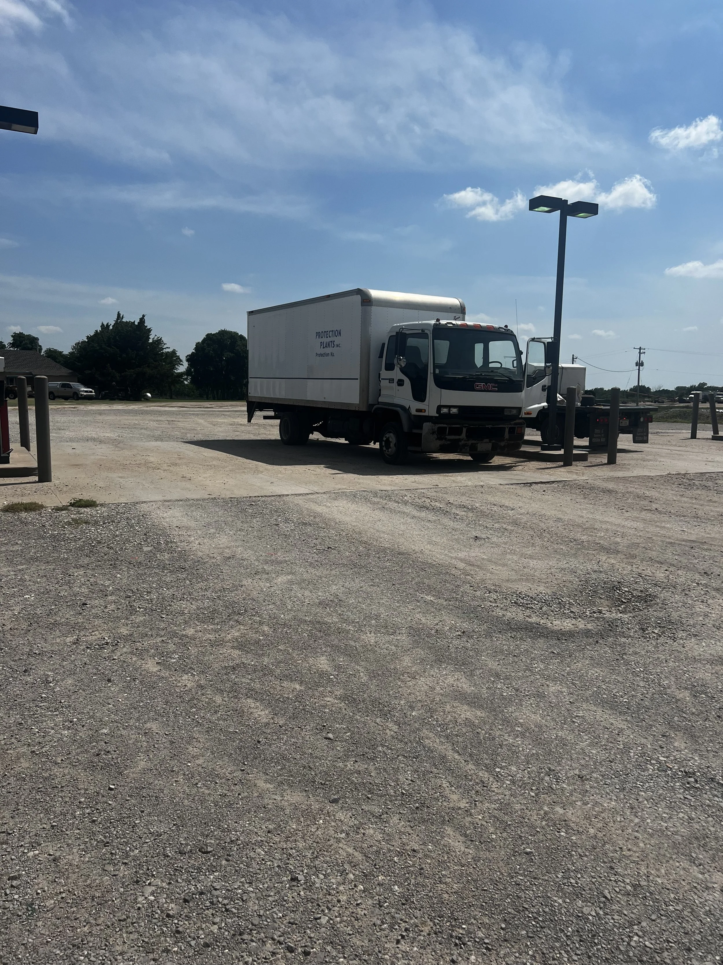 A white GMC box truck parked in an open gravel lot under a blue sky with scattered clouds, surrounded by a few trees and utility poles.