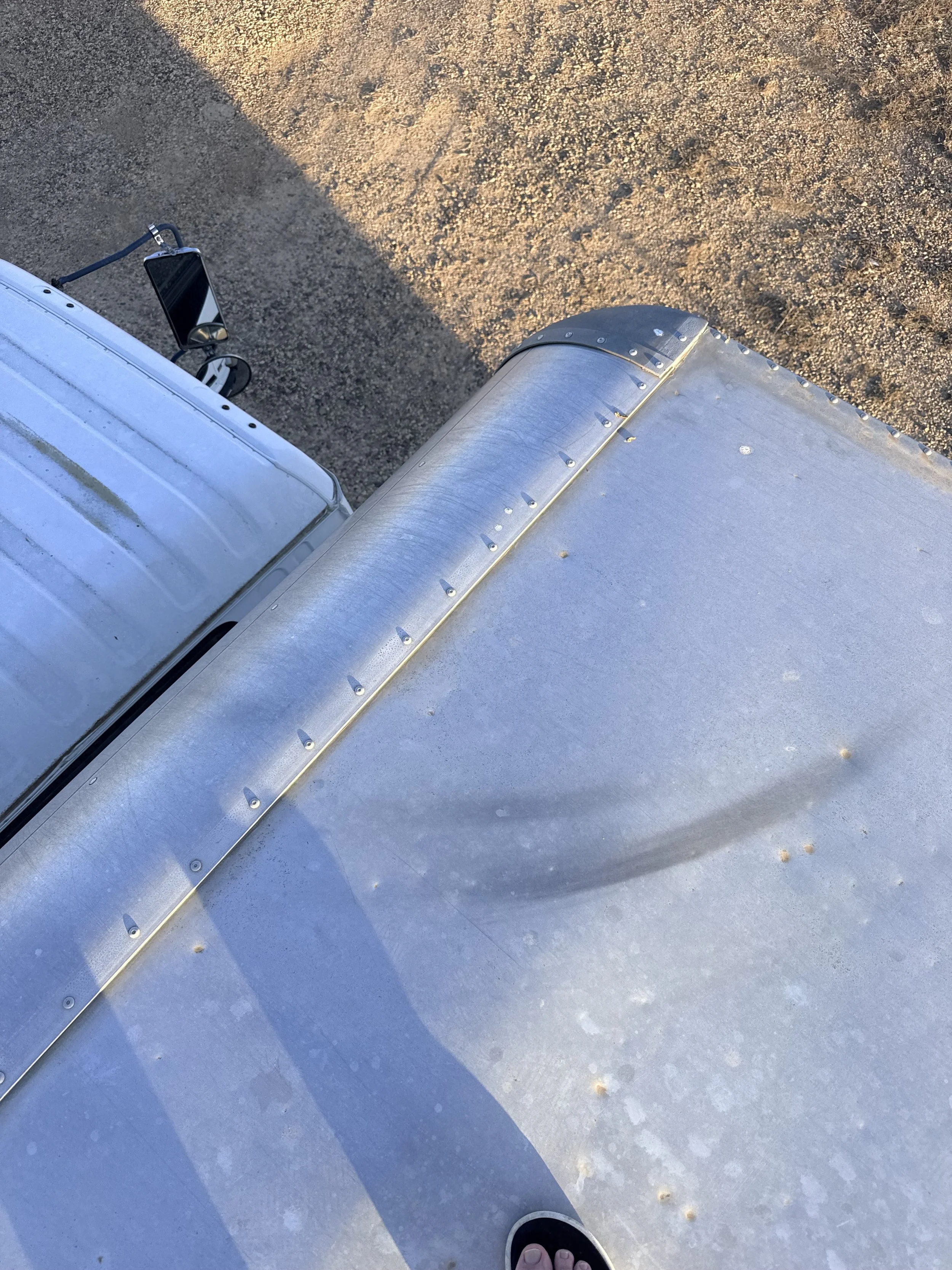 Top-down view of a person's foot in a flip-flop on a concrete surface with a metallic object, possibly part of a vehicle or machinery, and a mirror with a phone or device attached to it.