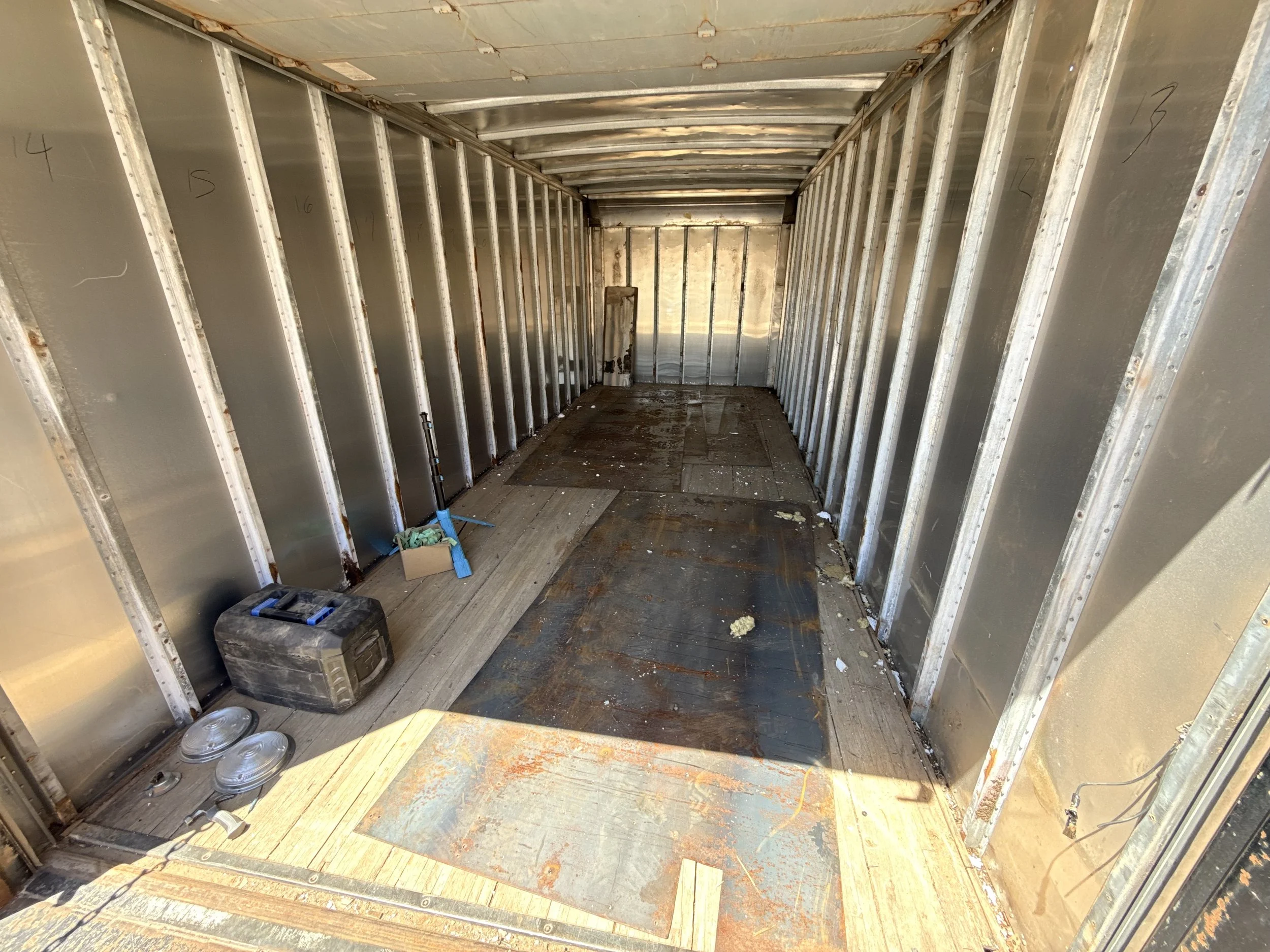 Empty interior of a small room under construction with metal wall studs, a wooden floor, and construction tools and materials on the floor.