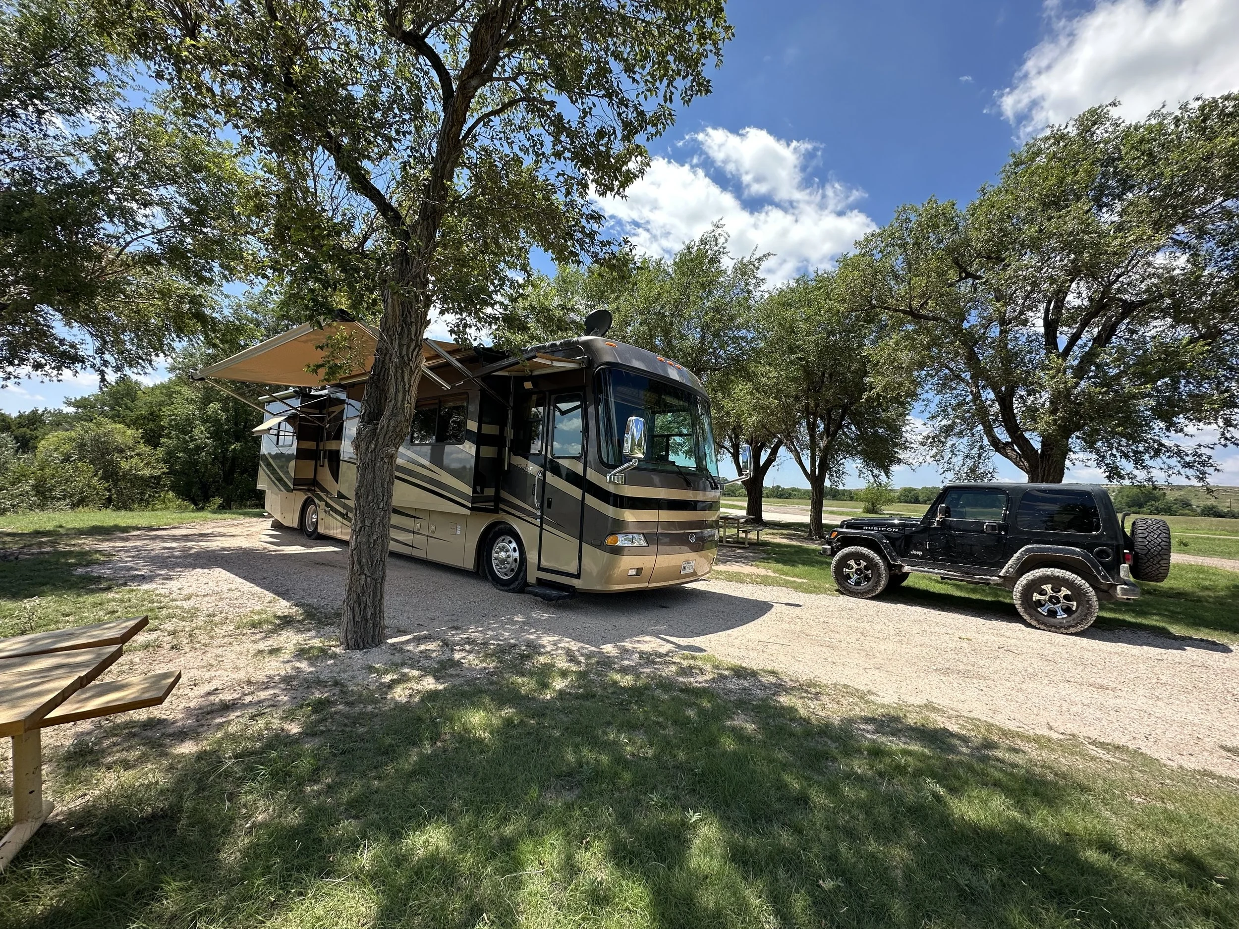 A recreational vehicle (RV) parked under trees on a partly cloudy day, with a pickup truck parked nearby and wooden furniture partially visible in the foreground.