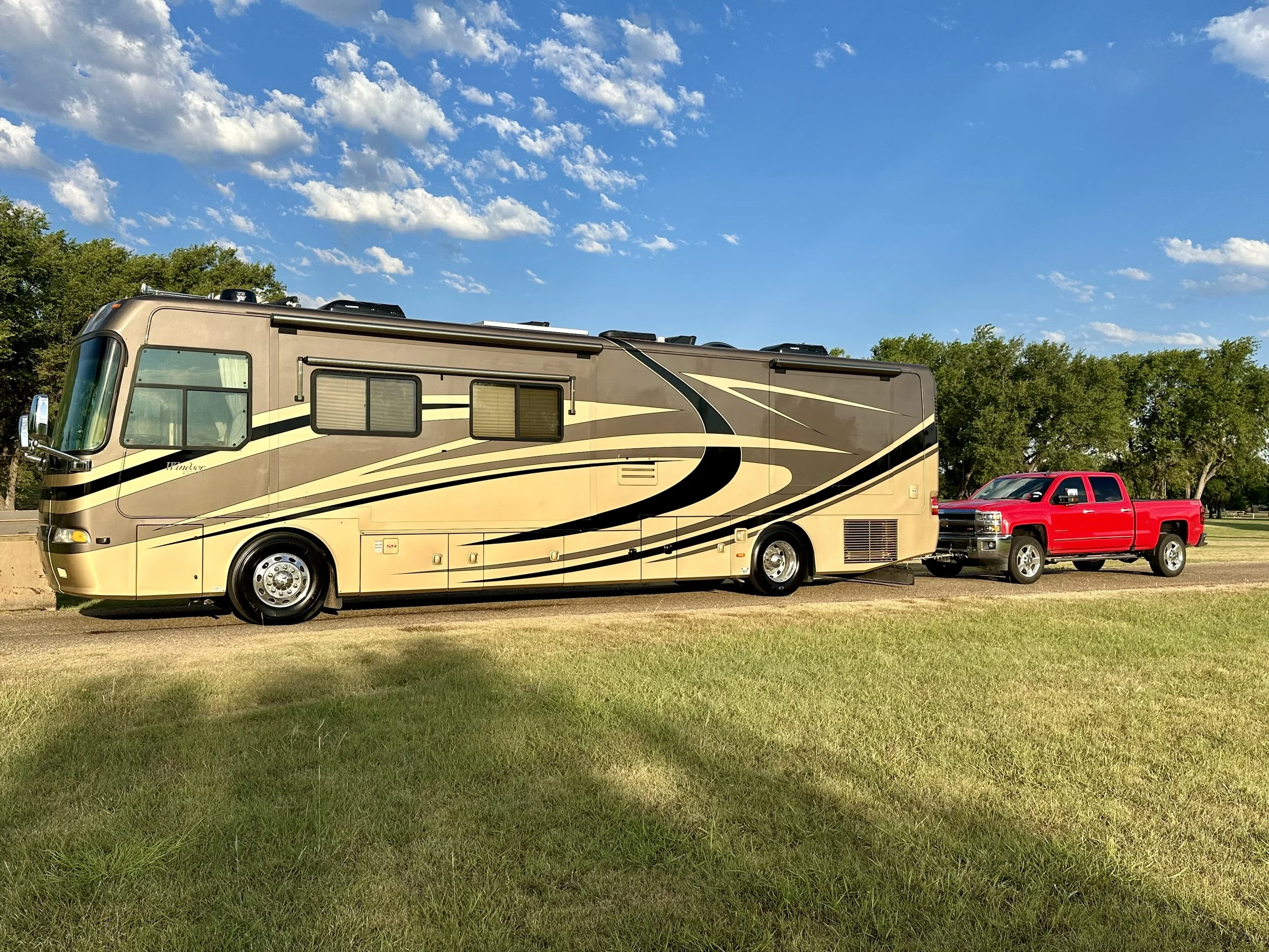 A beige and brown motorhome and a red pickup truck parked on grass with a backdrop of green trees and a partly cloudy blue sky.