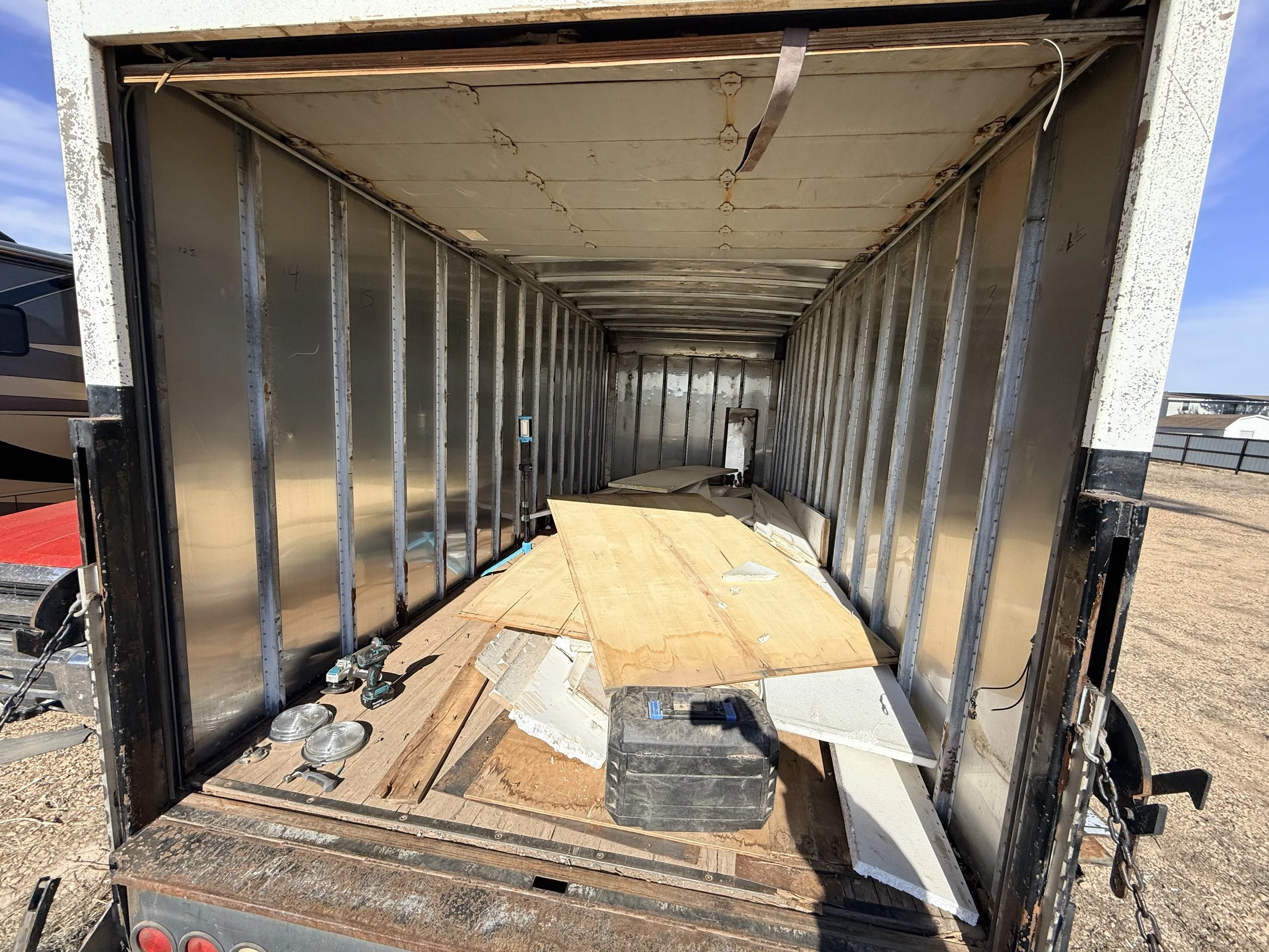 The interior of an empty cargo truck with metal walls and wooden flooring, construction tools and materials scattered on the floor.