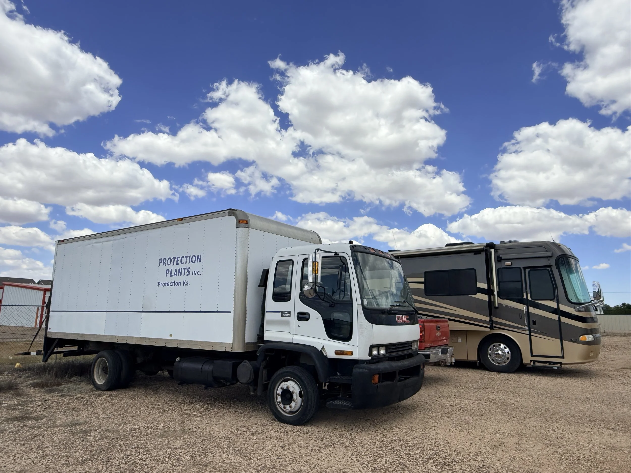 A white GMC delivery truck with a blank cargo box parked on a gravel lot, with a motorhome or RV beside it, under a bright blue sky with scattered fluffy white clouds.