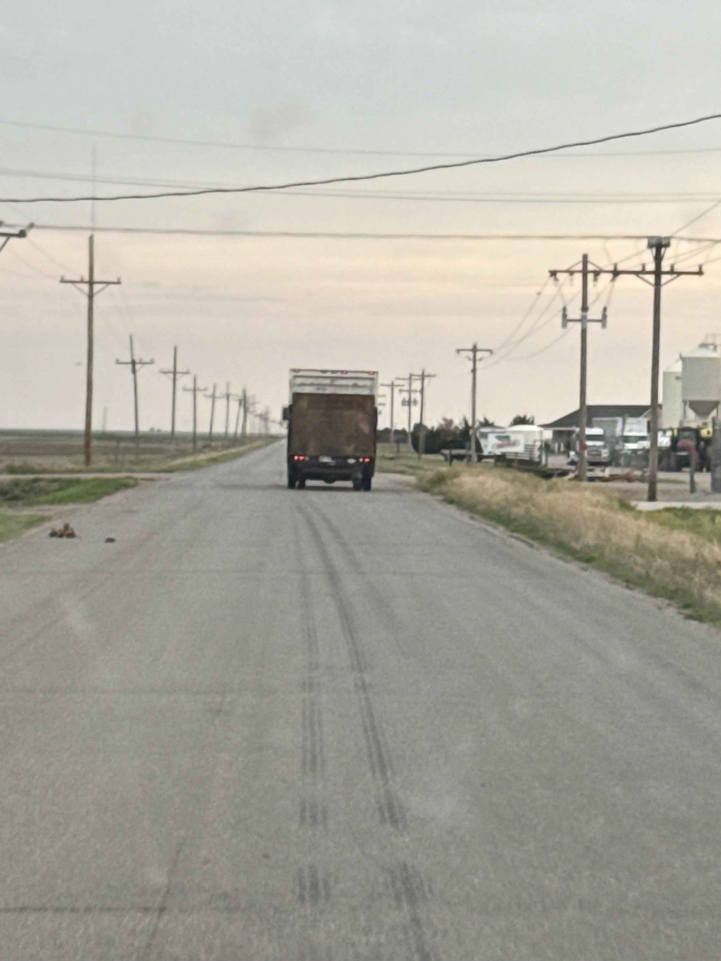 A rural road with power lines on both sides, a truck on the road, and buildings and other vehicles nearby, all under an overcast sky.