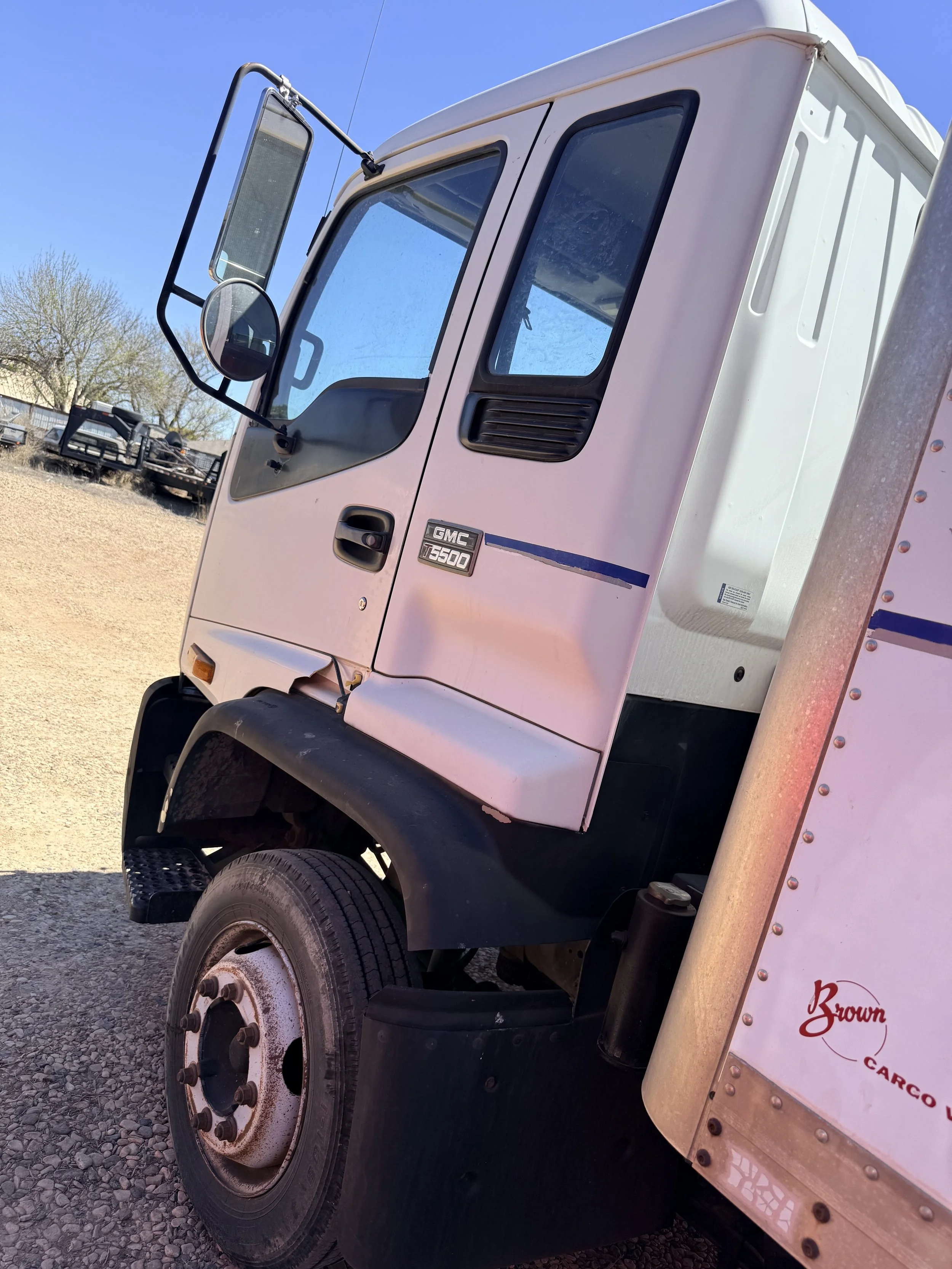 Close-up of a pink GMC T-5000 delivery truck with a truck cab, side mirror, and tire visible, parked on gravel under a clear blue sky, with some trees and other trucks in the background.