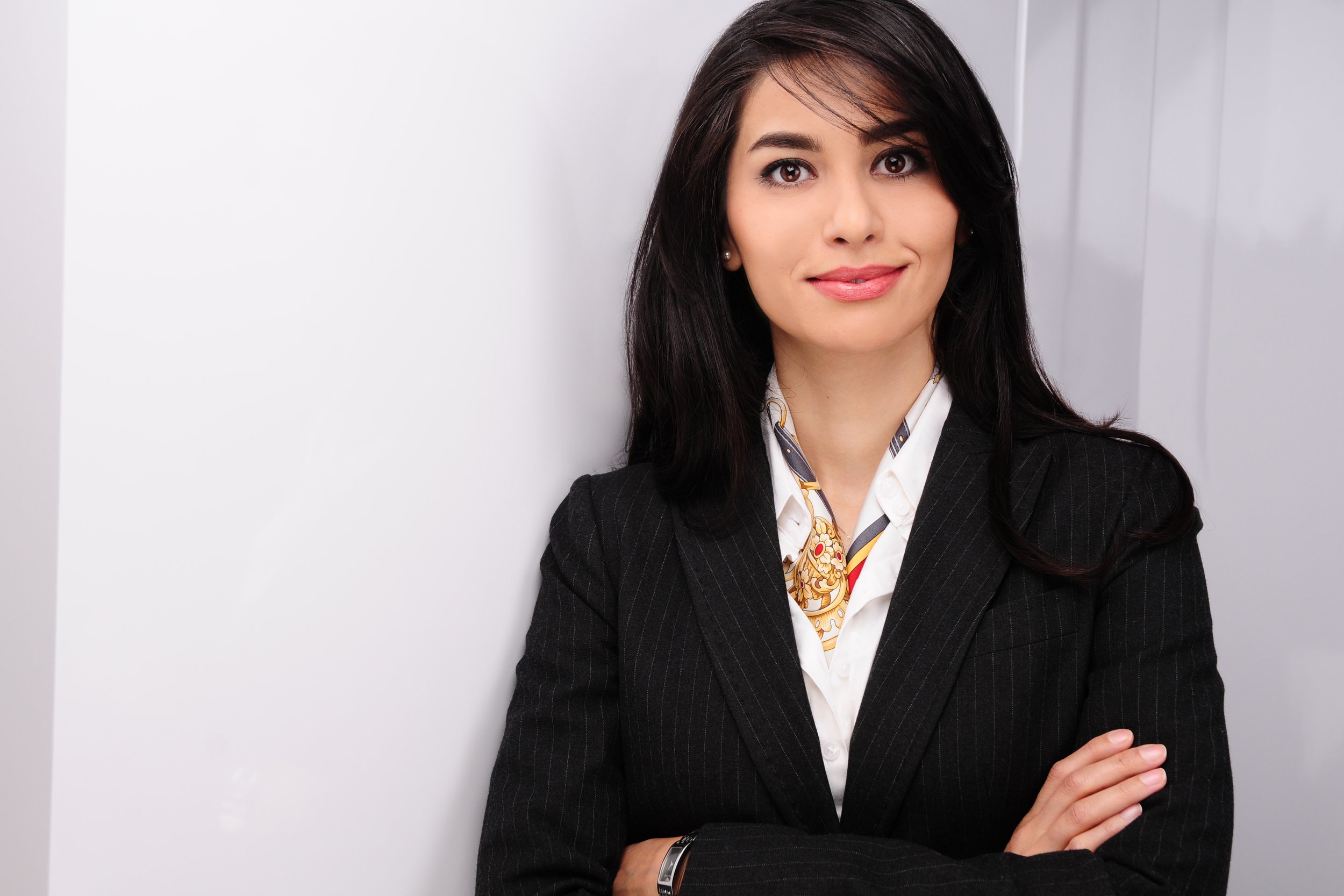 A woman with dark shoulder-length hair, wearing a black blazer and white shirt with a patterned scarf, standing against a light-colored background.