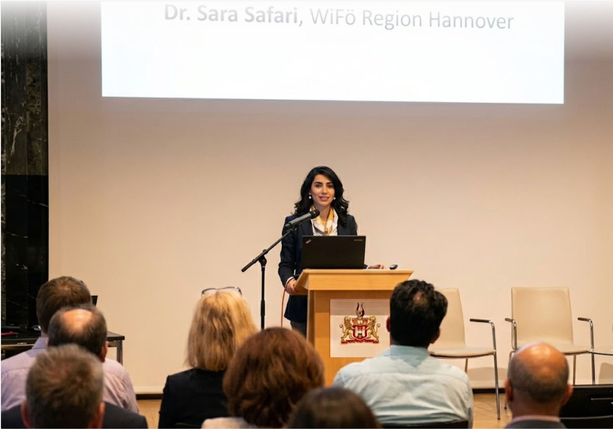 A woman with dark hair and a dark suit stands at a podium, giving a presentation in a conference room filled with seated attendees. A large screen behind her displays text that reads, 'Dr. Sara Safari, WiFo Region Hannover.'