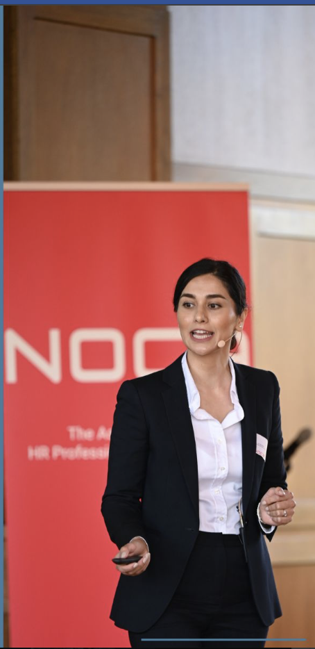 A woman in a dark business suit giving a presentation in front of a red backdrop with white text.