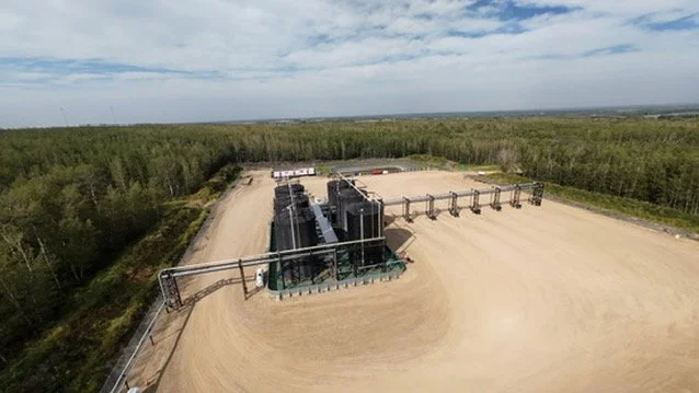 Aerial view of an industrial site in a rural area with large black tanks and pipes, surrounded by dirt roads and a forest in the background.