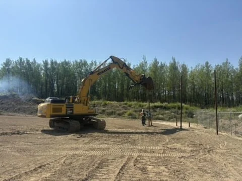 A yellow excavator on a construction site with a person nearby, trees in the background, and a clear blue sky.