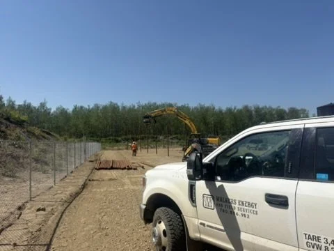 A construction site with a white utility truck in the foreground, a digger in the background, and a worker wearing an orange vest and helmet.