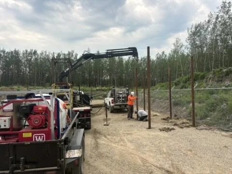 Workers installing a tall fence with wooden posts alongside a dirt road in a rural area, with trees and cloudy sky in the background.