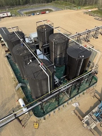 An aerial view of an industrial site with four large black storage tanks connected by metal pipes, surrounded by a safety fence, with a dirt area and a racetrack in the background.