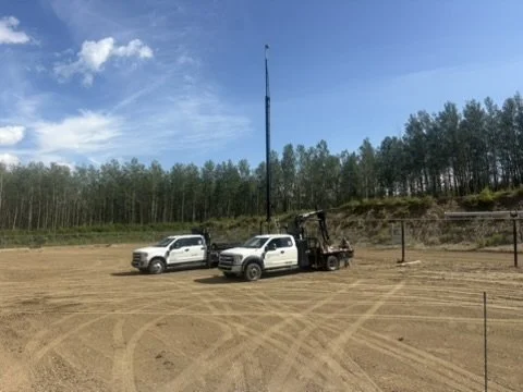 A dirt area with three white trucks, one with a crane, two trees, and a background of a forest under a blue sky with some clouds.
