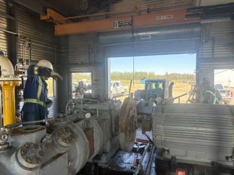 Workers in protective gear operate industrial equipment inside a building with large open doors, overlooking a parking lot and field outside.