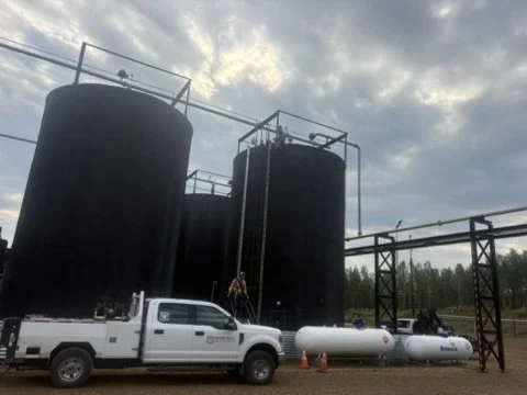 Two large black industrial storage tanks with a truck and smaller white tanks beneath them, set outdoors under a cloudy sky.