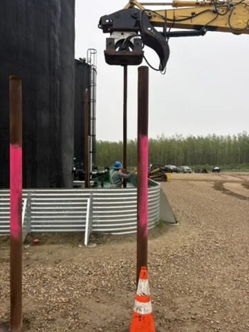 A construction site with a crane lifting a metal pole, which is secured by a traffic cone at the base. There is a large black storage tank and a building in the background, with a worker wearing a blue helmet nearby.