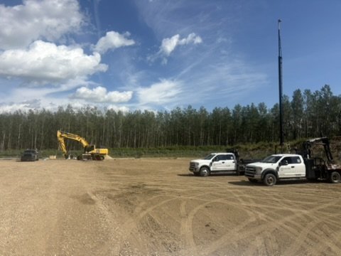 Construction site with excavator, several pickup trucks, and a flagpole against a backdrop of trees and blue sky with clouds.
