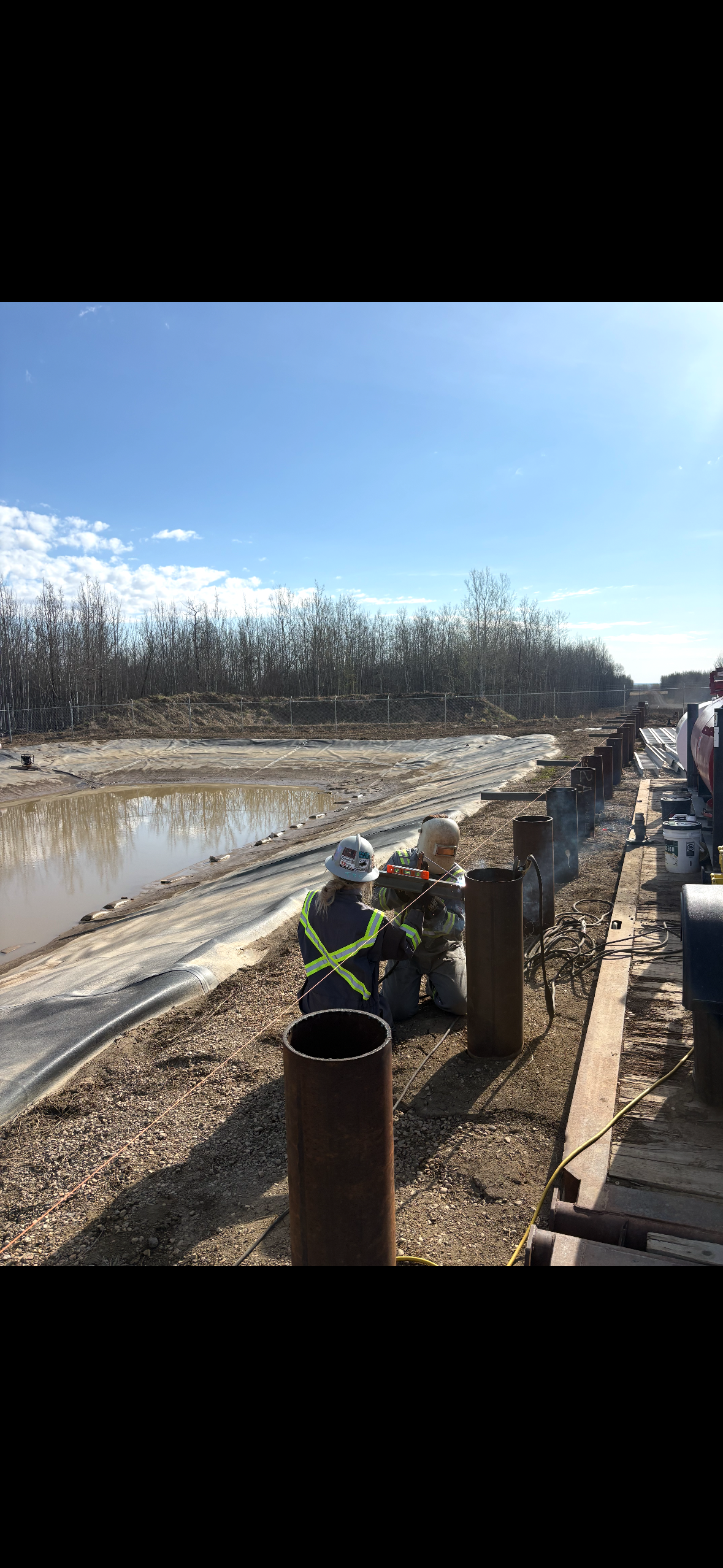 Two construction workers wearing helmets and reflective safety vests working near metal pipes, with a small pond and trees in the background on a sunny day.
