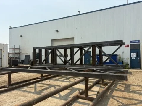 Metal framework structure outdoors near a building with pipes on the ground.