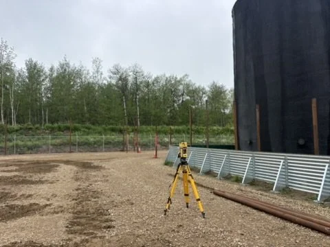 Construction site with a tripod-mounted surveying instrument, a large black tank or container, and metal pipes on the ground.