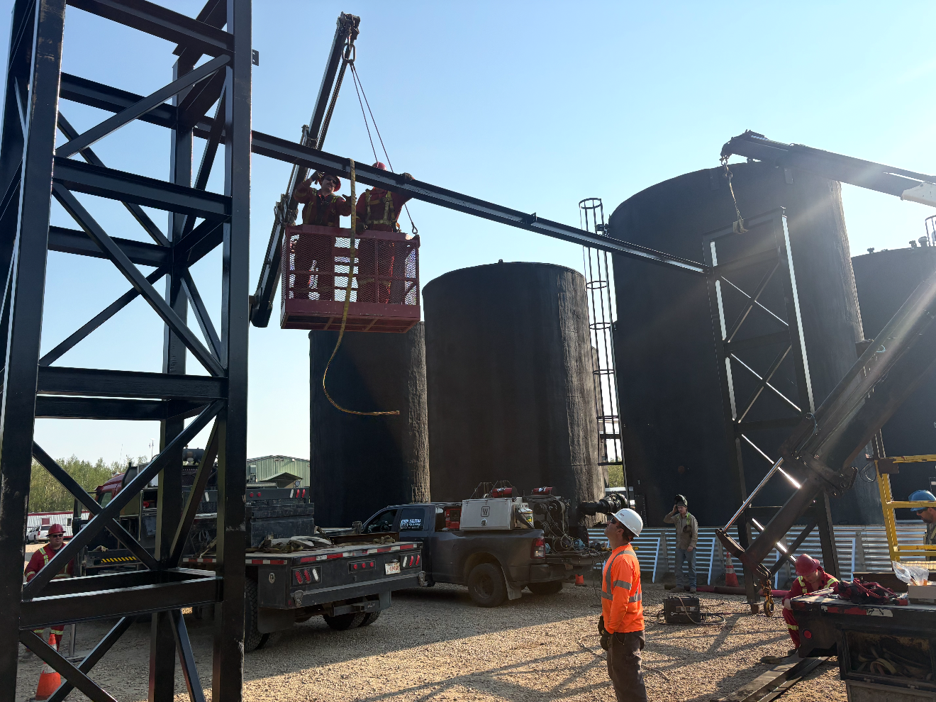 Workers in safety gear working at an industrial site with large black storage tanks, a crane lifting equipment, and various trucks and tools.