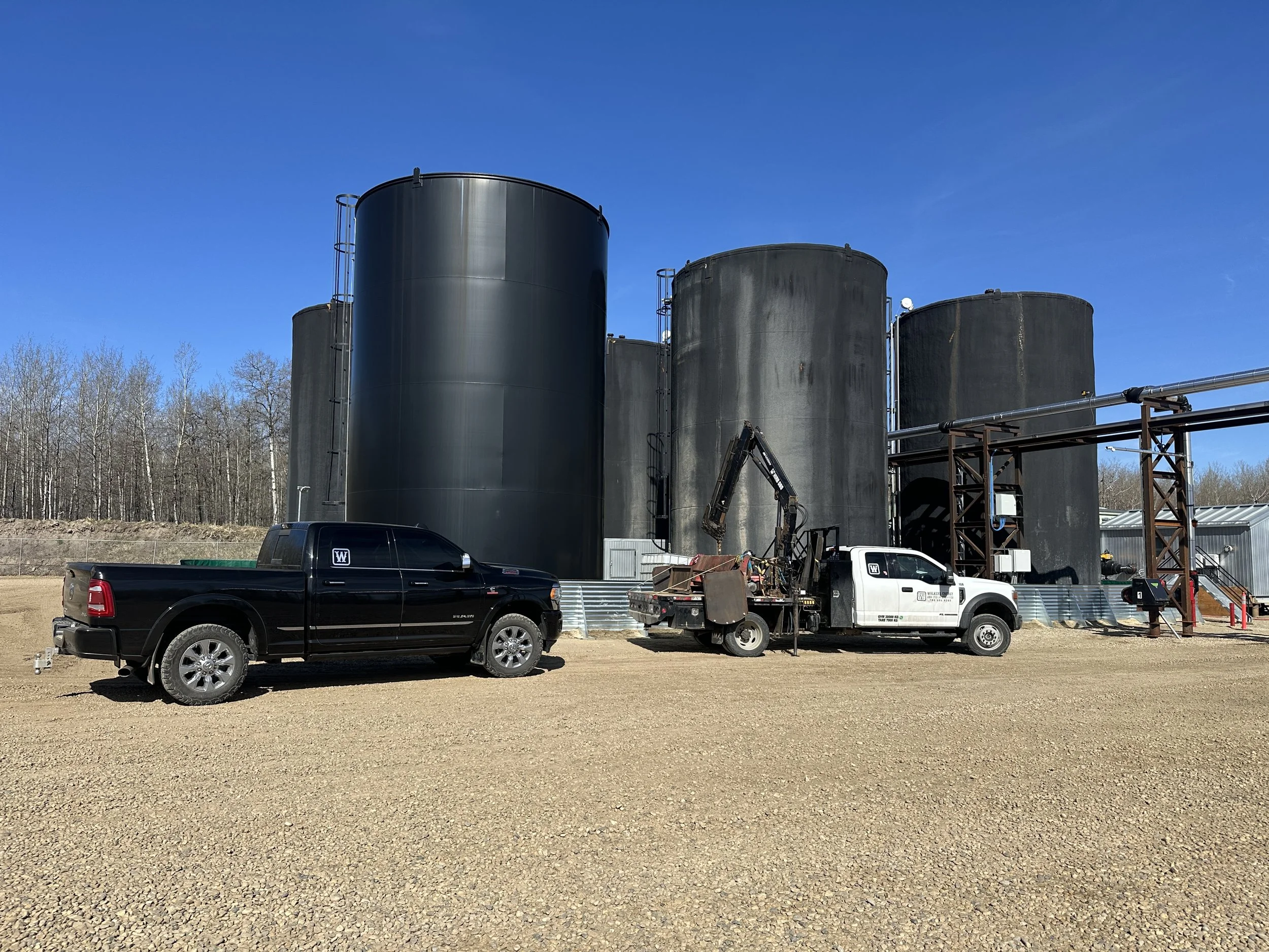 Large black industrial storage tanks with pipes, ladders, and a small crane in a construction site with two trucks parked in front and a clear blue sky