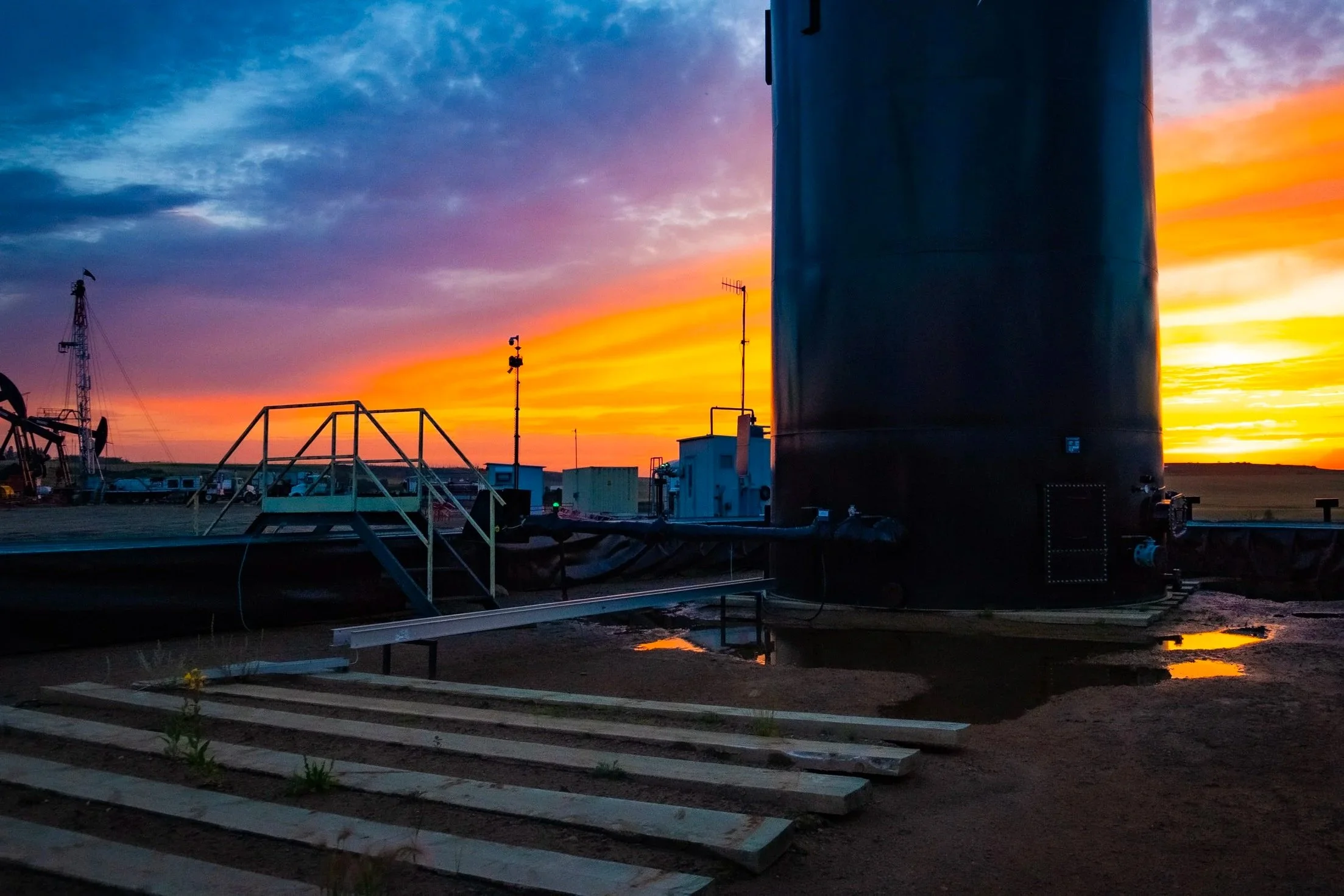 A landscape view of an industrial area at sunset with a large cylindrical tank and various small structures, water puddles on the ground, and a colorful sky with clouds and hues of orange, pink, and blue.