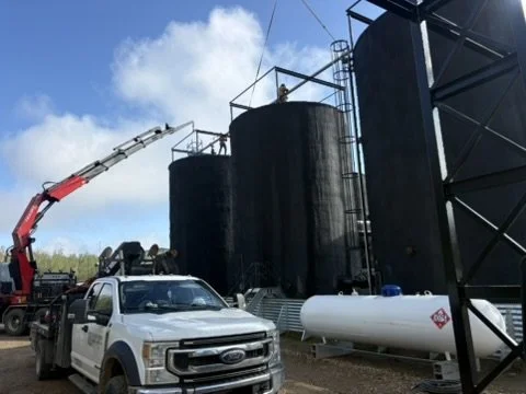 Firefighters spraying water from a fire truck's ladder to put out a fire on large black storage tanks at an industrial site.