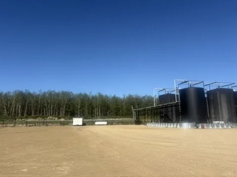 Industrial tanks and pipes overlooking a dirt field with a line of trees in the background under a clear blue sky.