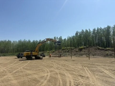 Construction site with a yellow excavator and workers installing fence posts on sandy terrain, with trees and clear blue sky in the background.