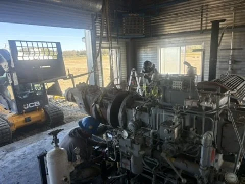 Two workers assembling or repairing a large industrial machine inside a workshop with windows, with a JCB forklift and outdoor landscape visible through the openings.