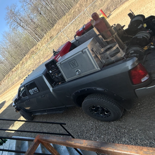 The back of a dark gray pickup truck loaded with firefighting equipment, including a fire extinguisher, metal storage box, and hoses, parked outdoors on gravel with a chain-link fence and trees in the background.