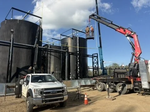 Two workers on a lift inspecting or repairing large black industrial tanks at an outdoor site, with a crane truck and a white pickup truck nearby, and an orange safety cone on the ground.