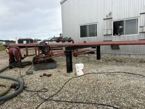 Pipeline system with red pipes, valves, and hoses outside a metal building with gravel ground.