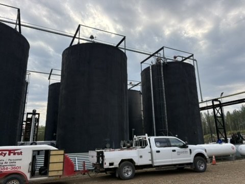 Four large black storage tanks with metal railings on top, a white pickup truck, and an equipment trailer parked outdoors under a cloudy sky.