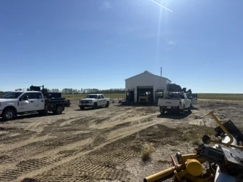Several pickup trucks parked in front of a small building on a dirt lot with tire tracks, under a clear blue sky.