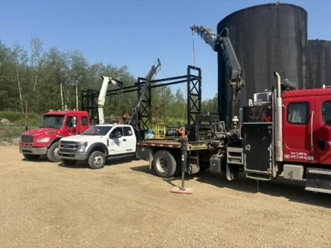 Three utility trucks, including one with a crane lifting equipment, parked on dirt ground with trees and blue sky in the background.