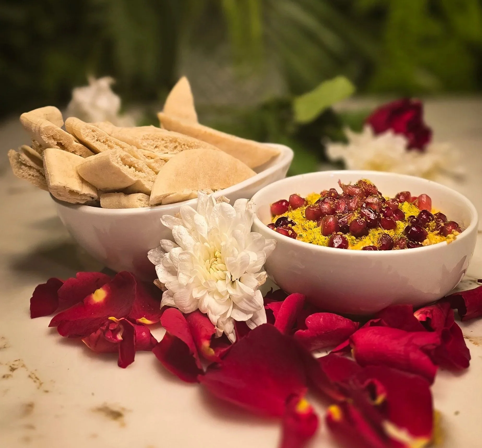 Close-up of a bowl of spiced houmous topped with pomegranate seeds garnished with yellow spices, surrounded by white and red flowers, on a white surface with rose petals scattered around. A popular menu item on ninety-seven bar Sidcup's small plates.