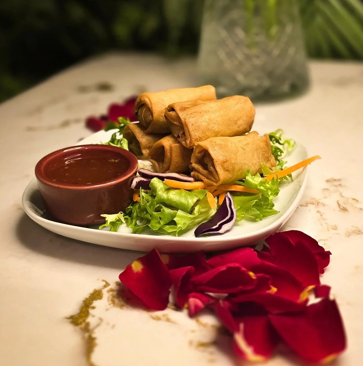 Plate of four spring rolls with a side of dipping sauce, arranged on a bed of mixed greens and vegetables, with red rose petals in the foreground. Popular vegetarian option at ninety seven bar Sidcup.