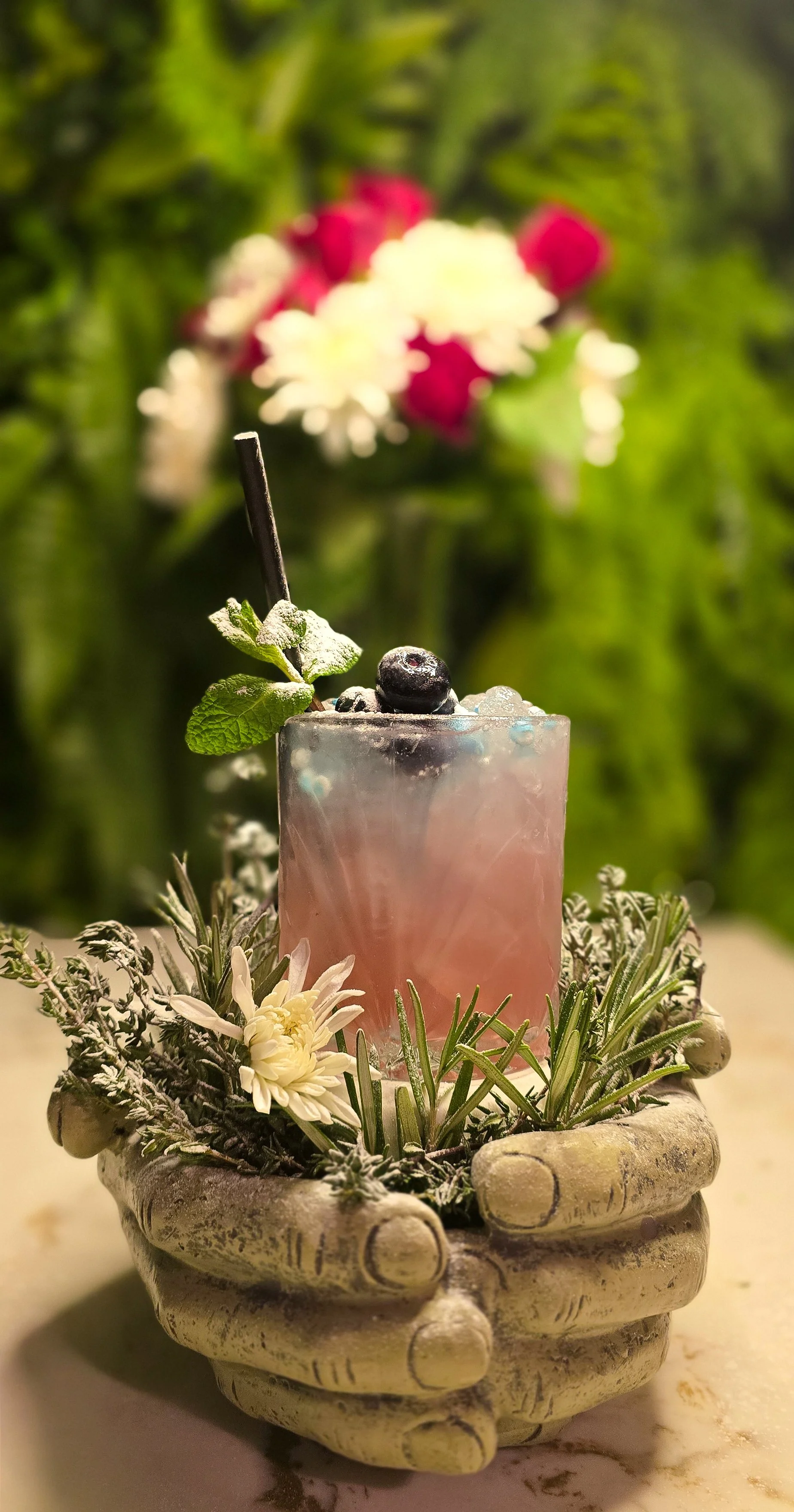 A pink bubblegum cocktail garnished with blueberries, a mint leaf, and a black straw in a frosted glass, surrounded by greenery, flowers, and stone pot. Signature cocktails at ninety seven bar Sidcup