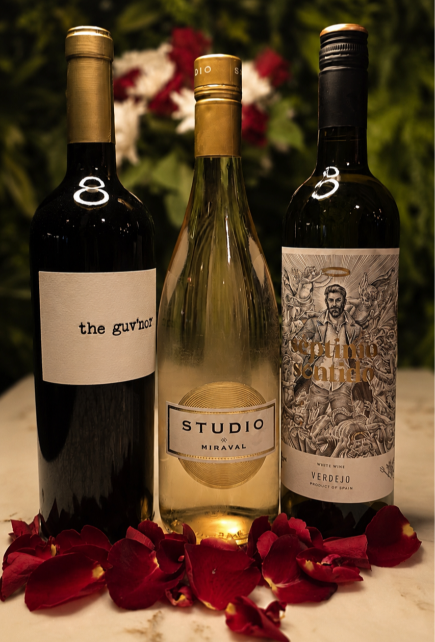 Three bottles of wine with red rose petals at the base, set against a blurred background of flowers and greenery, with warm lighting. Atmospheric wine at ninety seven bar and lounge Sidcup Kent.