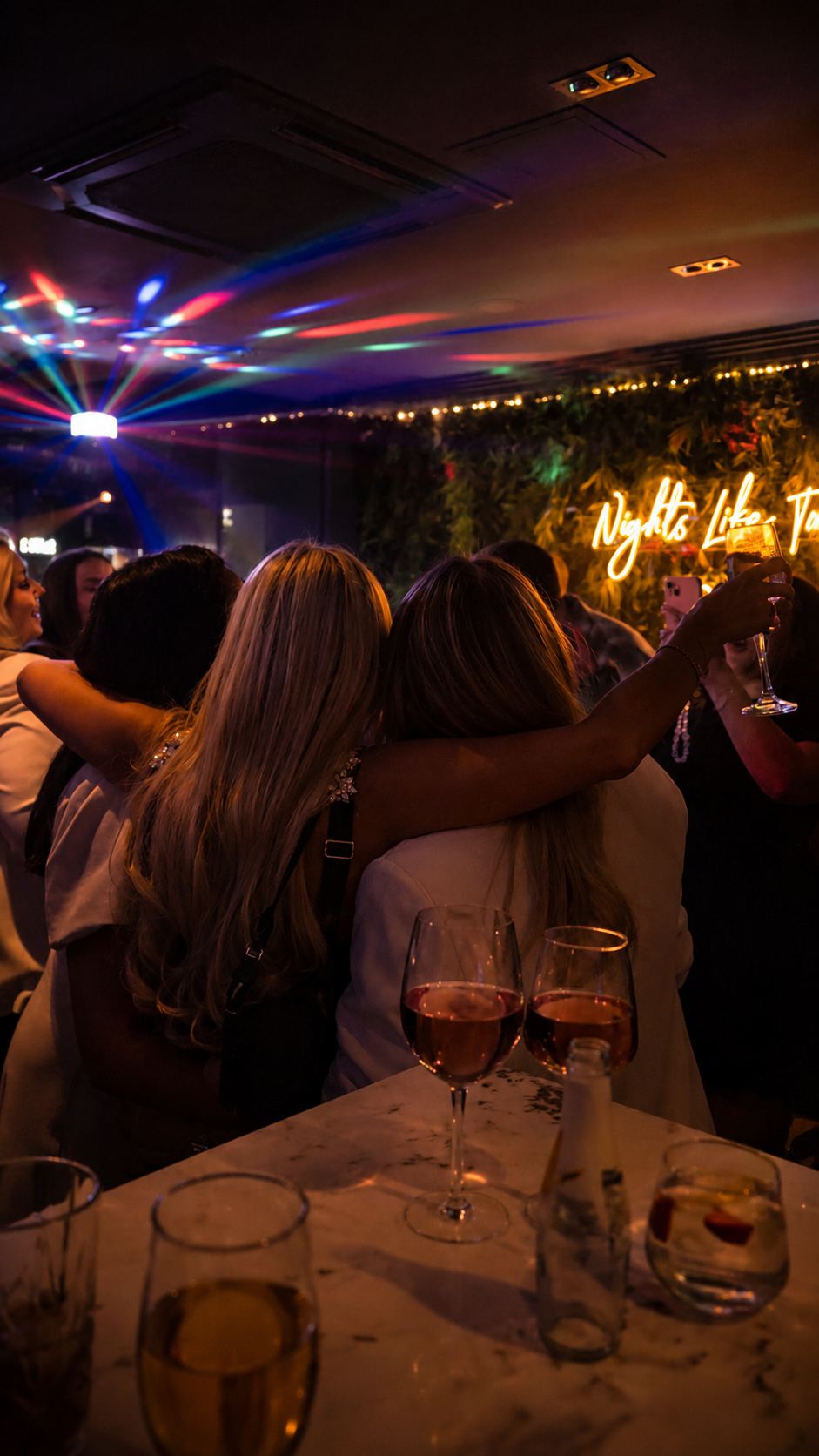 People hugging and celebrating at a party with colorful lights, glasses of wine, and a neon sign that says 'Nights Like This @ 97' on a decorated wall.
