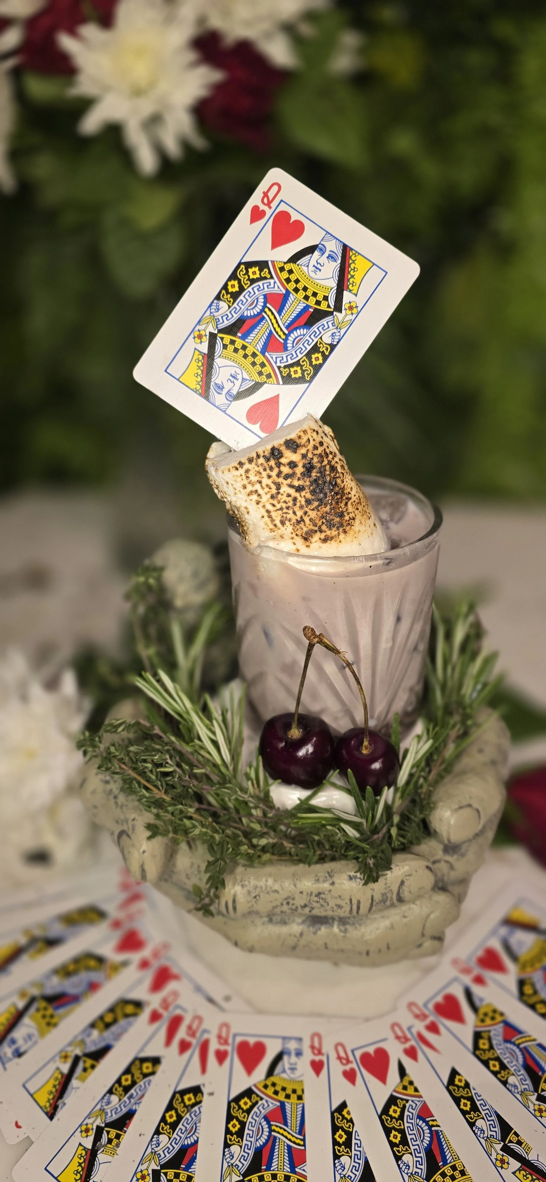 A marshmallow toasted with a card of the Queen of Hearts tops a luxury cocktail , surrounded by cherries and greenery in a decorative container, with playing cards laid out in a circle at 97 bar Sidcup .