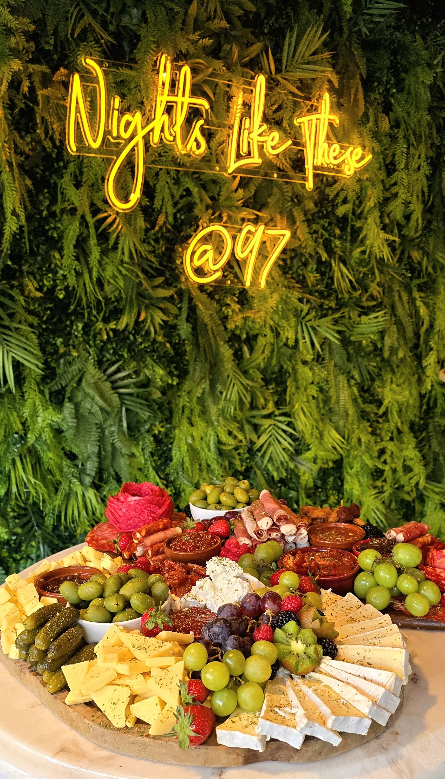 A cheese and fruit platter with grapes, strawberries, blackberries, and various cheeses, presented on a round wooden board. In the background, there is a lush green leafy wall. A neon sign above reads "Nights like these @ 97" in cursive yellow font.
